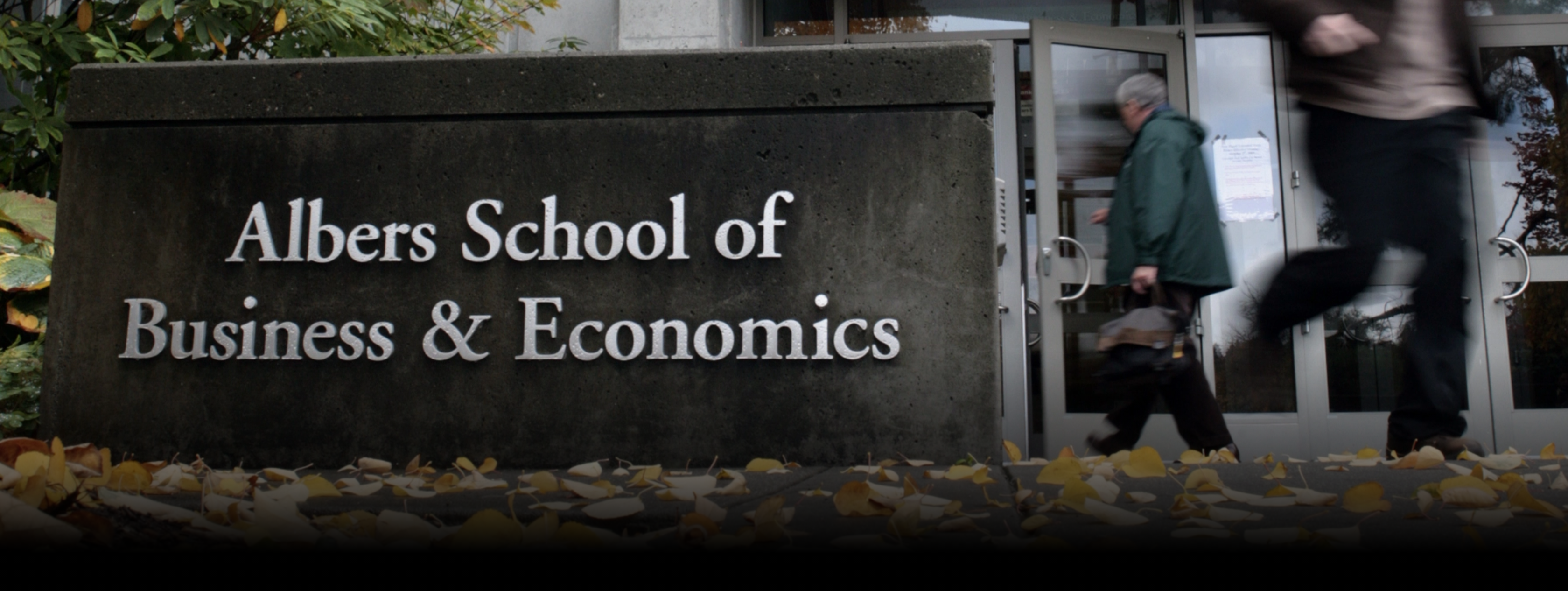 Sign reading Albers School of Business & Economics with fallen leaves and people walking past