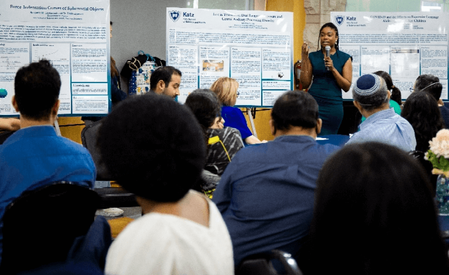 Woman with microphone addressing room full of students and presenting on easels