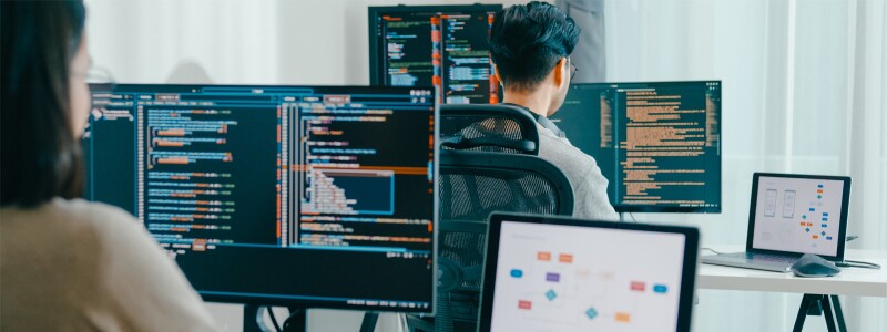 Two workers sit at desks in front of computer screens with data in the displays.