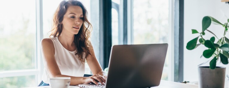 Woman with wavy hair sits in her home working on laptop