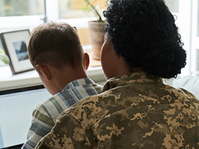 Military mother sits at dining room table with child on her lap while working on a laptop