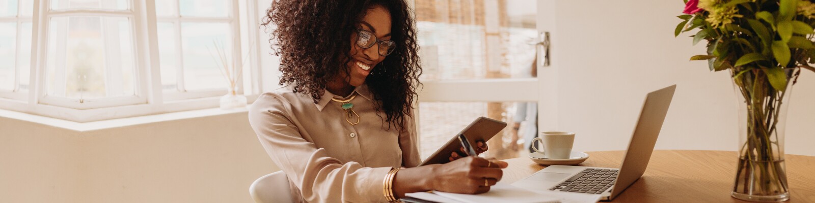 Woman with glasses holds a tablet while working on her laptop