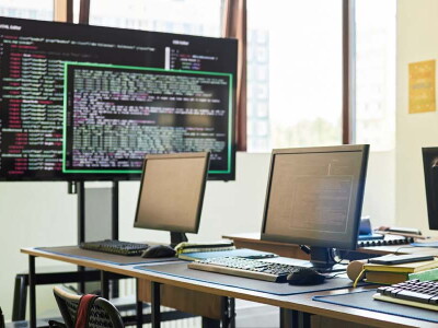 A sunny classroom is filled with various computers and screens displaying code.