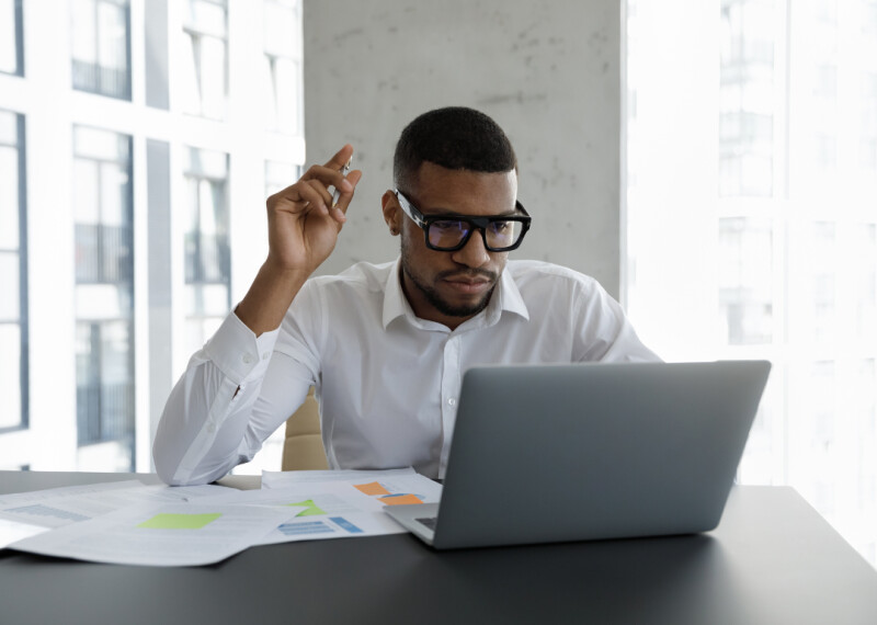 Man sits at table looking at lap top screen and holding pen aloft in his hand