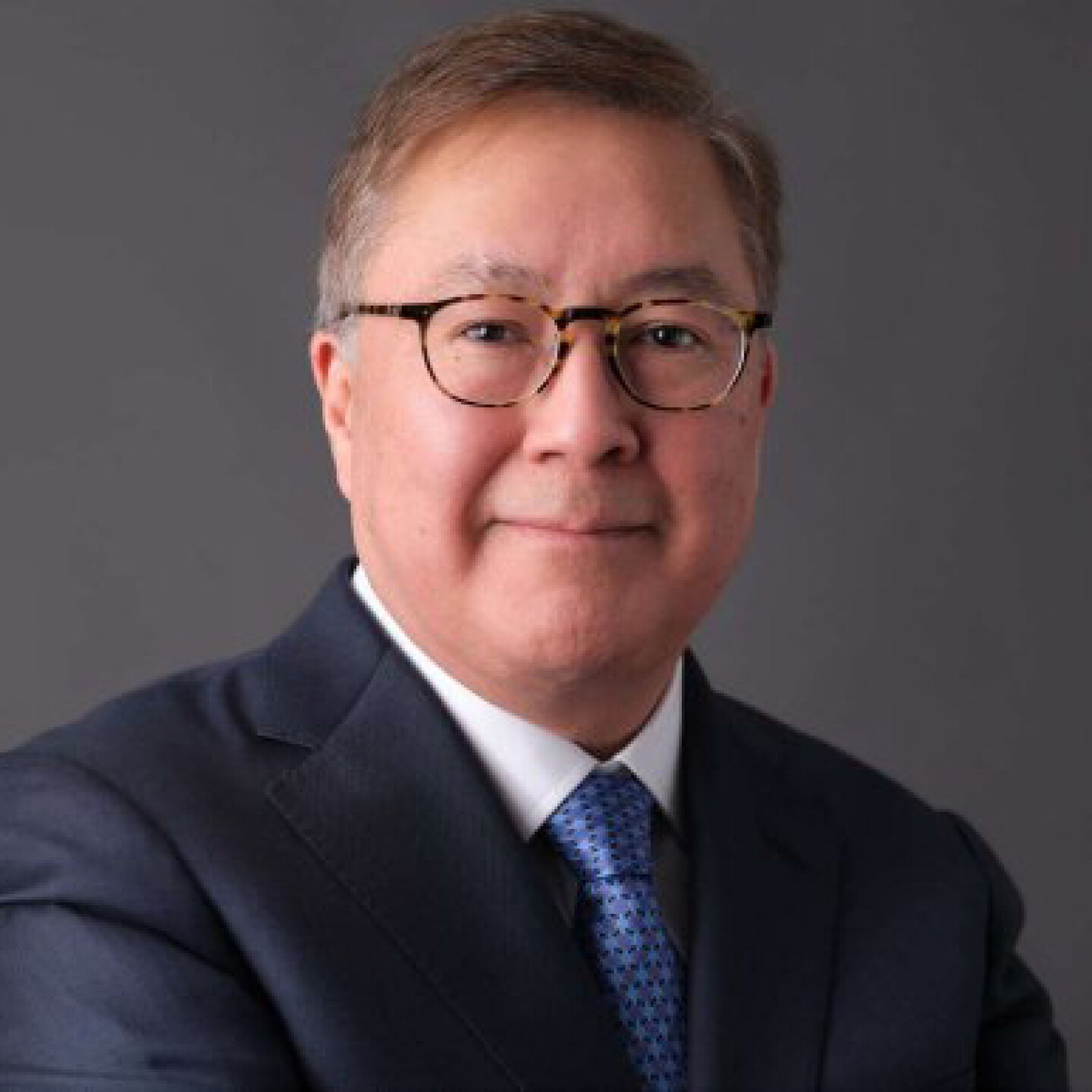Professional headshot of a man wearing glasses, a dark suit and blue tie against a gray background.