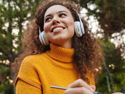 woman-in-yellow-turtleneck-wearing-headphones-writing-in-notebook-in-front-of-tree