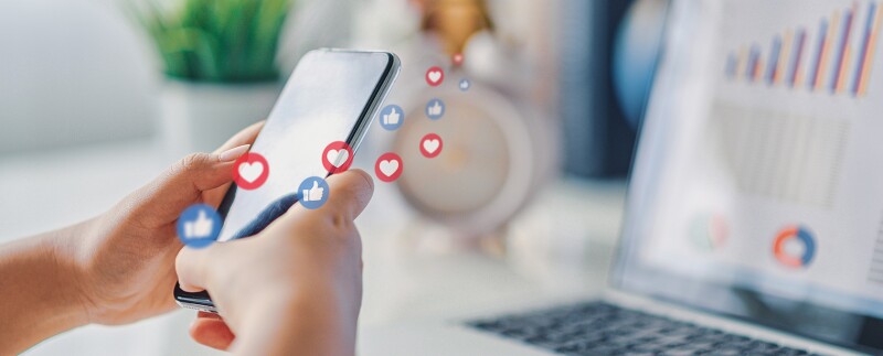 Hands using social media on a smart phone, laptop in the background with charts and graphs