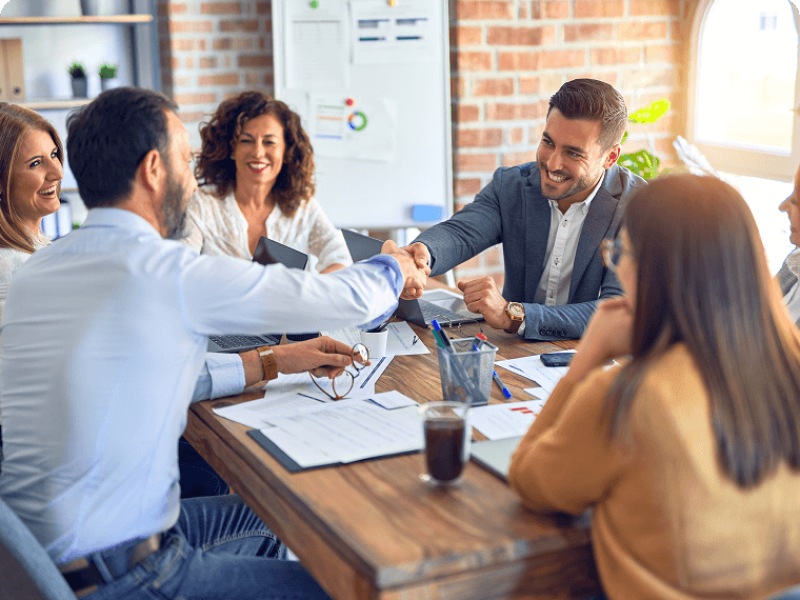 Two men shaking hands over a meeting room table with colleagues