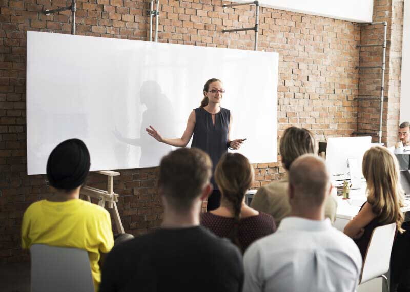 Young female professor standing in front of a white board speaking to a group of students.
