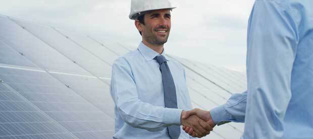 Two businessmen, one in a hard-hat, shake hands in front of a bank of solar panels.