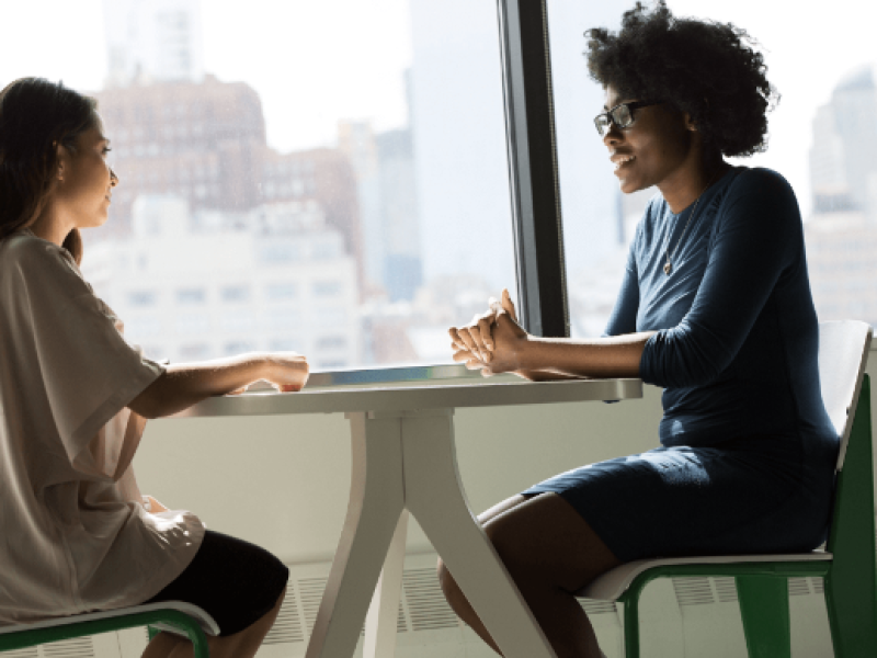 Two women seated at table in front of large window having a conversation