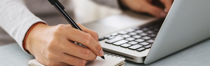 A person's left hand types on a laptop while the right hand jots notes in a notebook