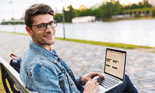 Man in Jean Jacket with glasses and laptop sitting on bench across from water