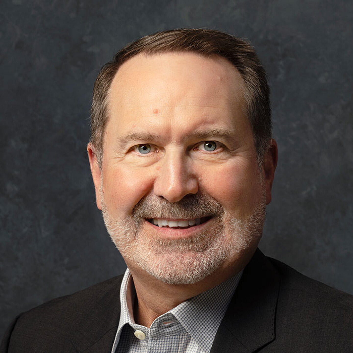 Smiling man with a gray beard wearing a dark blazer and checkered shirt against a dark gray background.