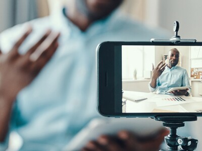Man sitting at desk films himself with a cell phone camera.