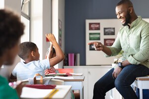 Smiling african american young male teacher gesturing on african-american elementary boy at desk.