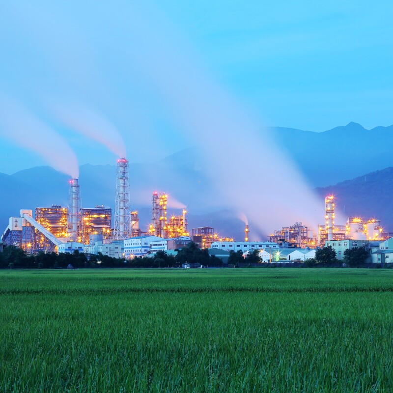 View of a factory in the middle of a green farmland in the early morning twilight ~ Factory pipes polluting air in a silent morning, a serious environmental issue