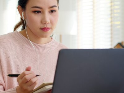 Asian female student wearing earphone while listening to digital online course development program.