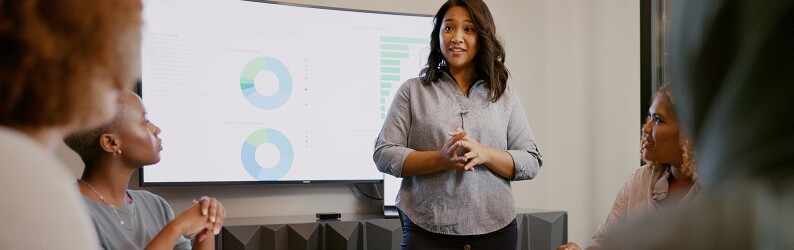 Woman stands in front of a group of her peers, presenting information depicted on a screen behind her.