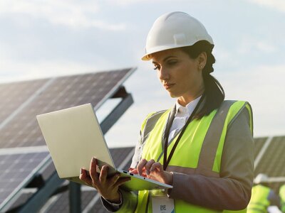 female engineer wearing hardhat standing near solar panels looking at laptop