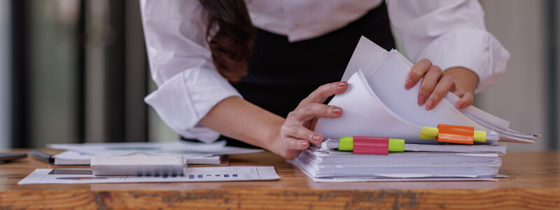 Close up shot of a woman's hands flipping through a thick stack of papers on a wooden desktop.