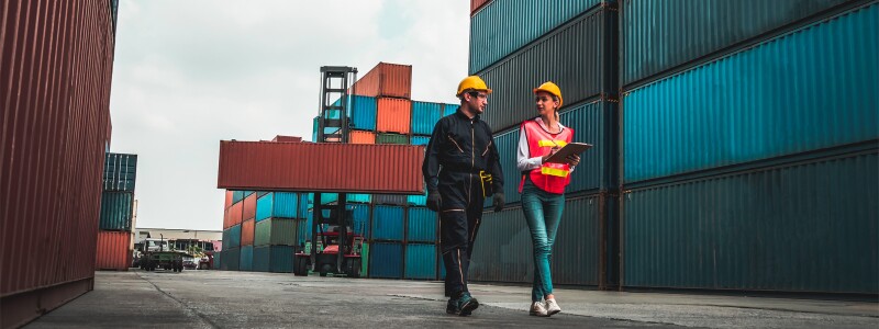 A man and a woman wearing hard hats walk through a work area filled with shipping crates.