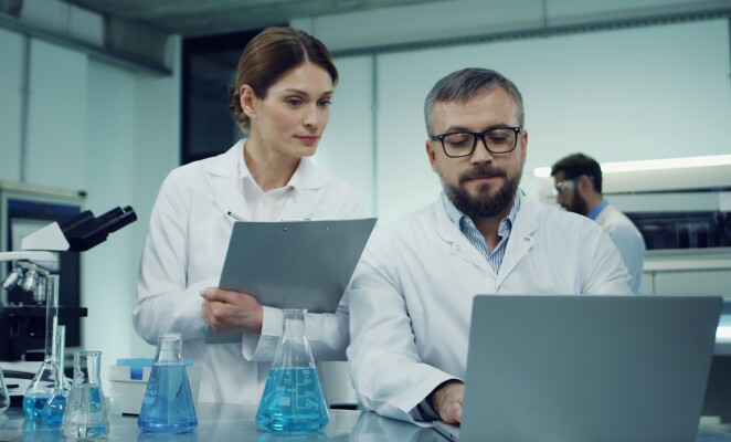 male and female scientists in the white robes working in a lab at a laptop computer doing research and taking notes