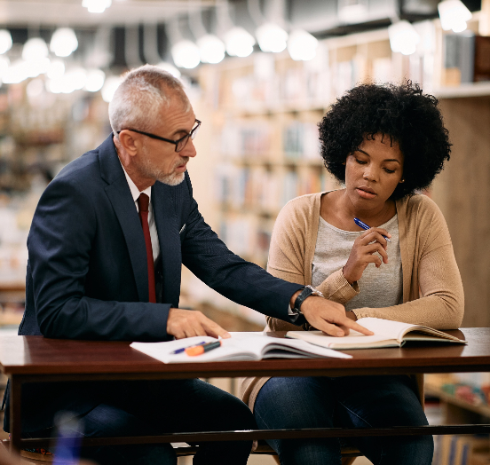Teacher and student stationed at a workspace within a library. The professor is explaining aspects of the students coursework to her.