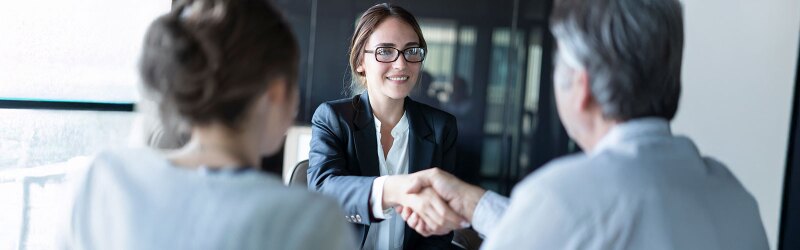 A woman in glasses and a suit smiles, shaking hands with someone across a table, potentially getting the job.