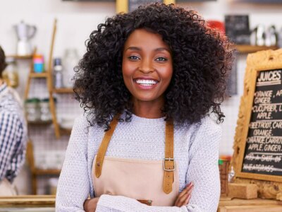 Smiling young African American female barista standing in her cafe.