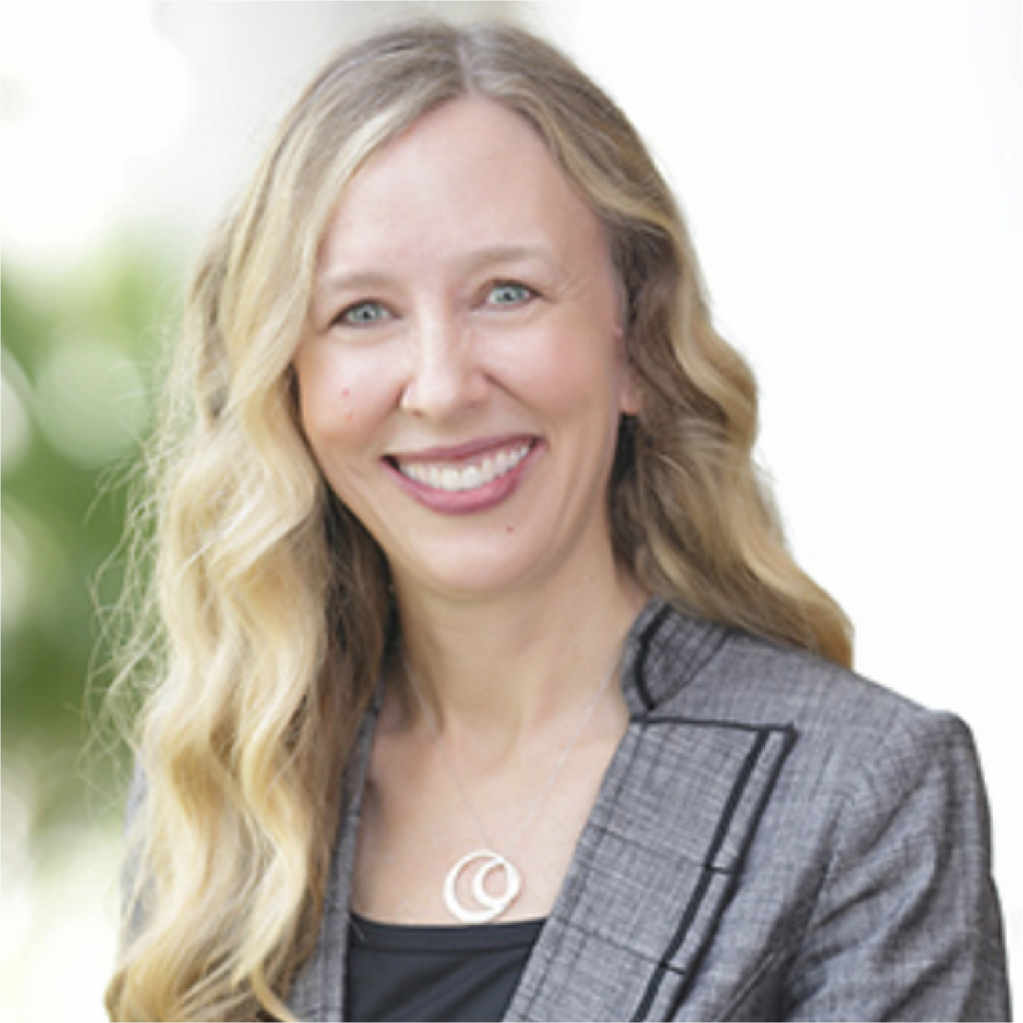 Headshot of a Beth Clarke with long blonde hair, wearing a gray blazer and necklace, against a softly blurred background.