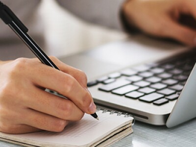 A person's left hand types on a laptop while the right hand jots notes in a notebook
