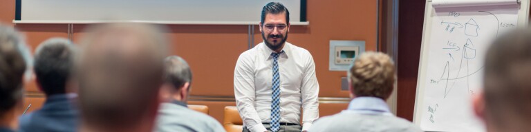 Professional in white button up shirt and tie sits in front of an audience of people