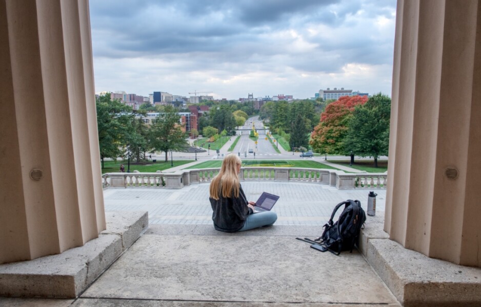 Female student sitting on steps in front of campus building with backpack and laptop