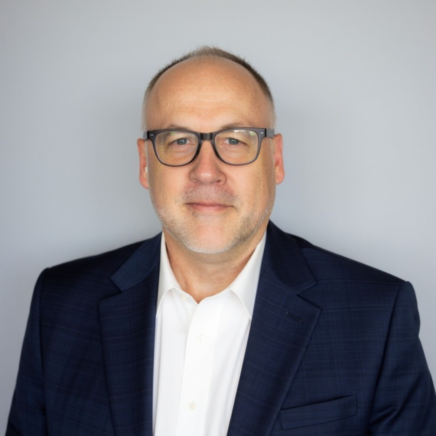 Professional headshot of a man with glasses, wearing a navy blazer and white shirt, against a light gray background.