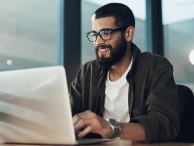 Man in cardigan and T-shirt smiling while he works on laptop on table
