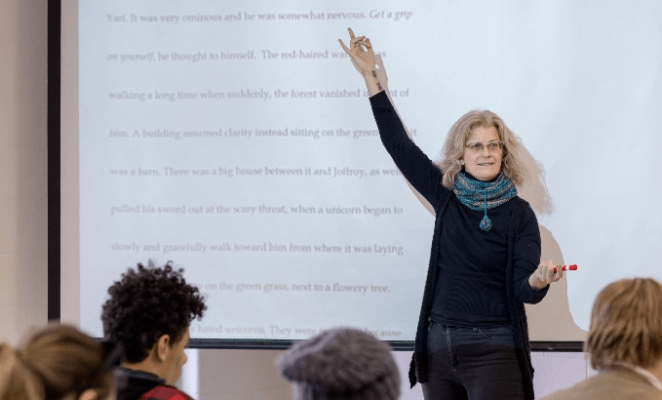 Female instructor at front of class gesturing toward literature displayed on a projection screen