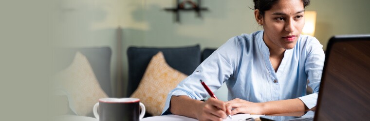 Woman wearing light blue tunic holds pen while looking at a laptop screen in living room