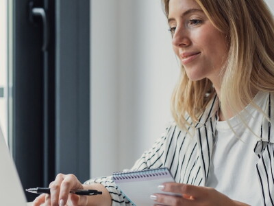 Woman working on a laptop with a notepad in her hand