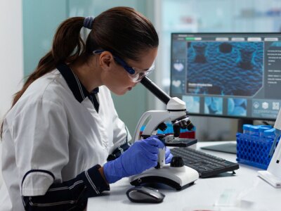 Female bioengineer is analyzing biomedical virus sample using medical microscope working in pharmacology hospital laboratory.