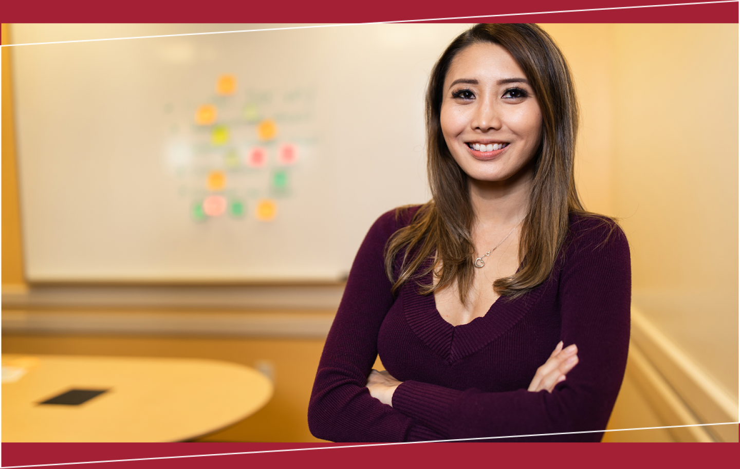 Smiling woman with a whiteboard that has colorful sticky notes on it blurred in the background.