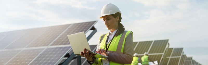 female engineer wearing hardhat standing near solar panels looking at laptop