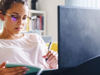 Young woman wearing glasses makes notes in notebook while working with laptop on her lap