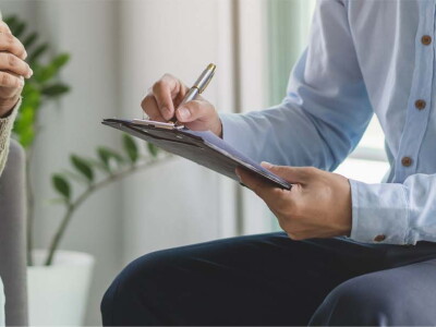 Close-up image of a man with a clipboard and pen sitting across from a woman.