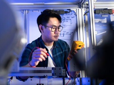 Electrical engineer using a digital multimeter to check voltage on an industrial machine, with a robotic machine in the foreground.