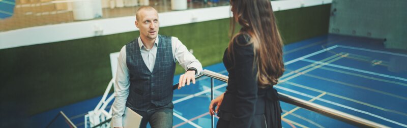 Male sports scout discusses recent scouting trip findings with female director in an indoor court.