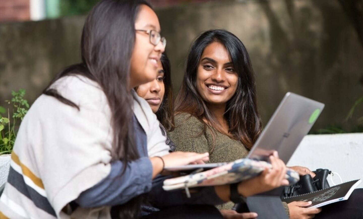 Three woman seated on bench smiling and looking at laptop