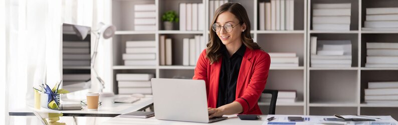 smiling woman in red blazer sitting in white office space typing on laptop