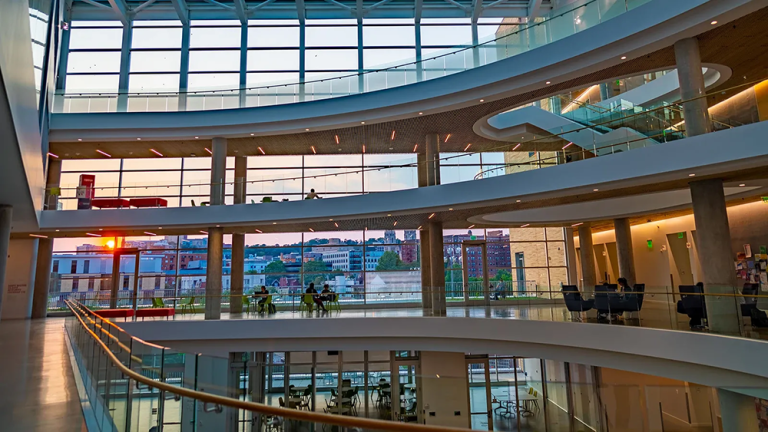 Modern multi-story university atrium with curved balconies, floor-to-ceiling windows, and sunset city views through expansive glass walls.
