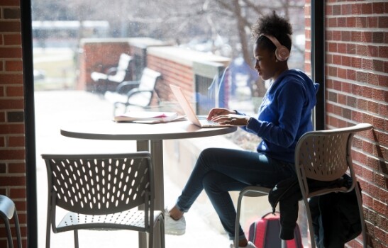 Student with headphones in front of a window working on laptop